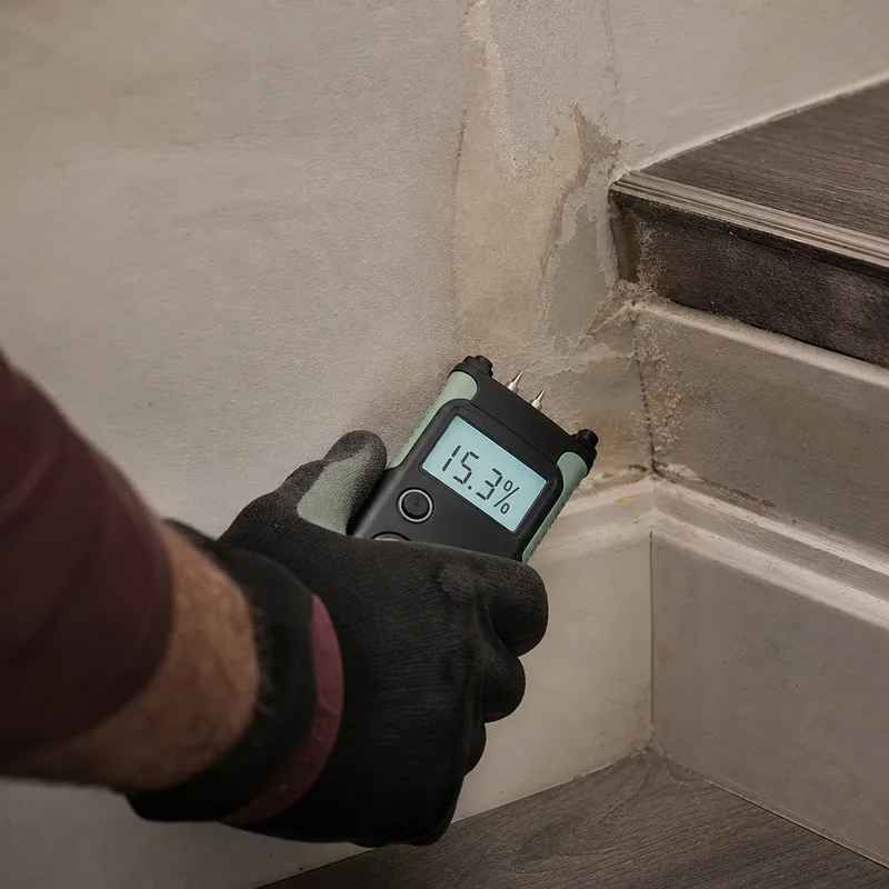 Moisture meter reading on baseboard and drywall at the bottom of a basement stairwell