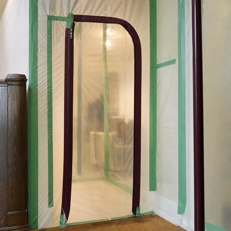 Plastic sheeting containment wall with a zippered entry doorway and taped seams in a residential hallway